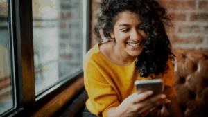 Smiling woman in yellow sweater using smartphone near window, representing professional mobile technology communication